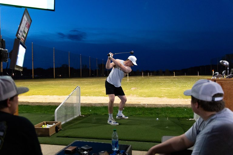 A group of people playing in a golf league at TopCityGolf at night under the bright lights, hitting golf balls with their clubs on the driving range